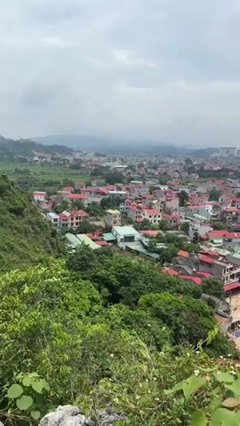 A panoramic view of a densely populated urban area with many houses featuring red tile roofs. The houses are of varying sizes and designs, with some appearing to be older and others newer. Green trees and vegetation are interspersed throughout the town, and a lush green hillside is in the foreground. In the distance, rolling hills are visible under a cloudy sky. A few larger buildings, possibly commercial or institutional, can be seen among the houses, and a tall communication tower is also visible in the far distance. The overall impression is of a vibrant, lived-in community nestled within a natural landscape.