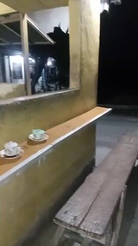 The video shows a close-up of a ledge with two tea cups and saucers on it. In the background, through a window or opening, there is a dimly lit room with shelves holding bottles and glasses. The overall environment appears to be outdoors or in a covered outdoor area, possibly a cafe or a home's veranda, with a rustic and somewhat worn aesthetic. The lighting is dim, suggesting it is evening or night. The camera is positioned to focus on the tea set, with the background slightly out of focus.