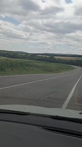 The video captures a view from inside a vehicle, looking forward and slightly to the right. The road ahead is paved and curves gently to the right. White lines mark the lanes. On the left side of the road, there is a field of green crops, possibly corn. Beyond this field, a line of trees separates it from rolling hills covered in a mix of fields and forests. The sky is partly cloudy with a bright, overcast sun. On the right side of the road, there is a white speed limit sign with the number 20 inside a red circle. The landscape appears rural and agricultural.