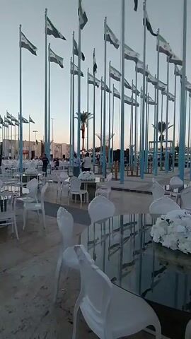The video depicts an outdoor event space set up with numerous round tables and white chairs, adorned with floral centerpieces. The background features a dense array of flagpoles, each flying the flag of Syria. People are seen milling around, some formally dressed, suggesting a possible celebration or gathering. A stage is also visible, implying the potential for performances or speeches. The setting appears to be at dusk or dawn, given the sky's soft lighting. The camera moves across the scene, offering various views of the setting and attendees.