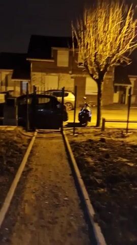 The camera is moved forward down a narrow dirt path flanked by two concrete edges. There is a dark car, a light-colored car, and a motorcycle parked behind a dark metal fence. The trees are bare. The houses in the background have lit windows. It is dark.