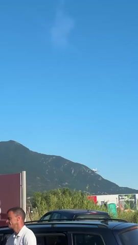 A static, vertical shot shows a clear blue sky with a faint white contrail above a distant green mountain range. A white airplane flies from right to left, positioned over the mountain range. Below the mountains, green vegetation and parts of buildings are visible, including a structure with a red roof. In the midground, a large maroon sign with the word "KORO" is partially visible. The immediate foreground features the roof of a dark vehicle with roof rails and the upper part of a man's head and shoulder in a white shirt, positioned on the left.