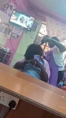 From the mirror's reflection in what appears to be a salon, a person in a white-and-purple uniform is using a hair dryer on a customer's hair. The customer, seen from the chest up, is holding a purple smartphone and looking at it. The salon has pink and green walls, and a television is playing. There are images of hairstyles on the wall. An electrical outlet is visible on the lower left corner of the mirror.