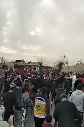 The video depicts a large crowd of people, primarily dressed in dark clothing, many with bicycles, gathered in what appears to be a public square or plaza. Most are wearing helmets. Several people are waving Syrian flags. Some individuals are standing on elevated structures, playing instruments, and using mobile phones to film the scene. The environment is overcast, and there are buildings and trees visible in the background. The crowd is chanting, suggesting a form of demonstration or rally.