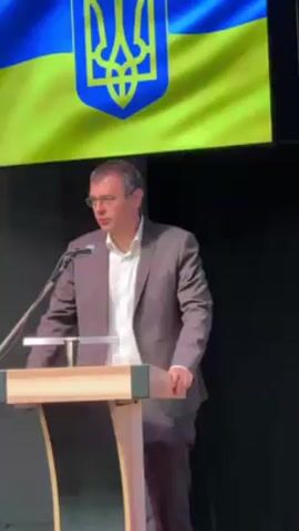 A man in a brown suit stands at a wooden podium giving a speech. He is wearing glasses and a white collared shirt. Behind him is a large Ukrainian flag. The background is black. A microphone is positioned in front of him on the podium. The podium has a simple, modern design with a flat top and slightly curved support.