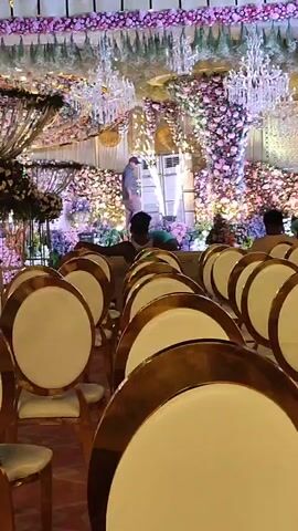 The video captures an elaborately decorated indoor event venue, likely a banquet hall, in Askihala, India. Rows of elegant white and gold chairs are neatly arranged, facing a grand stage or altar area. The entire space is lavishly adorned with extensive floral arrangements, predominantly featuring pink, purple, and white flowers, creating a vibrant and festive atmosphere. Several sparkling chandeliers hang from the ceiling, contributing to the opulent decor. In the background, near the stage, a few individuals, possibly event staff or organizers, are visible. One person in a light blue shirt walks across the stage, indicating ongoing preparations or final checks for an event. The venue appears to be set up for a formal occasion such as a wedding reception or a similar celebration, but it is currently empty of guests. No visible text is present.