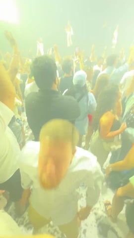 The video shows a crowded concert venue with many people, their arms raised, enjoying the music. Confetti is falling from the ceiling, and stage lights illuminate the performers, whose identity is obscured. The environment is dark, but the stage lights create a vibrant atmosphere. Some individuals are holding up their phones.