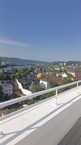 A vertical video pans across a rooftop balcony overlooking a city on a sunny day. The balcony has a white railing and gray composite decking. In the background, there are residential buildings, some with white facades and dark roofs, and others with warmer tones. Trees are scattered throughout the cityscape. Further in the distance, a body of water, possibly a river or lake, is visible, with bridges and more buildings on the opposite shore. The sky is clear and blue with a few wispy clouds. The camera movement is smooth and gradual, offering a panoramic view of the urban landscape.