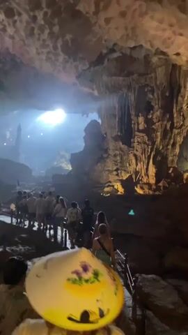 A group of tourists walk along a path inside a large cave. Sunlight streams in from an opening in the cave ceiling, illuminating the scene and creating dramatic lighting effects. The cave walls and ceiling are covered in stalactites and stalagmites, natural rock formations. Many of the tourists are wearing light-colored shirts and appear to be following a guide. In the foreground, a person wearing a conical hat is visible, with the hat's painted design partially obscuring the view of the cave. The path appears to be made of stone or concrete and has wooden railings.
