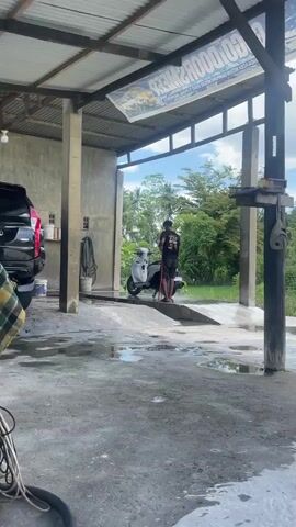 A young man in a black t-shirt and shorts is washing a white scooter at an outdoor car wash. He is using a red hose to spray water on the scooter. The scooter is parked on a ramp. There are palm trees and other greenery in the background. The car wash appears to be open-air, with a corrugated metal roof supported by concrete pillars. The ground is wet and there are puddles of water. A red car is partially visible on the left side of the frame.