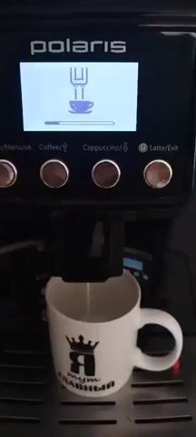 The video shows a close-up shot of a Polaris coffee maker in operation. The screen displays a graphic of a coffee machine brewing a cup of coffee, with a progress bar underneath indicating the brewing process. Below the screen are four buttons labeled "Menu/ok", "Coffee", "Cappuccino", and "Latte/Exit". The video focuses on the spout of the coffee maker, from which a white liquid, presumably milk or cream, is being dispensed into a white mug. The mug has a black crown graphic and Russian text that appears to say "Я тут главный" (I am the boss here). The liquid level in the mug rises as the coffee maker continues to dispense. The lighting is bright and appears to be from an overhead source.