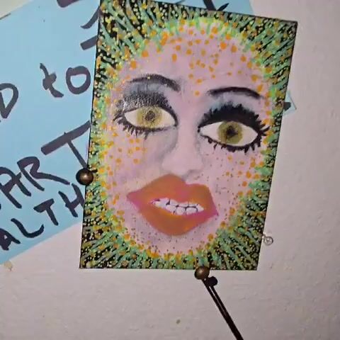 The video shows a close-up of a painting of a face, seemingly a woman's. The face has exaggerated features with large eyes, defined eyebrows, and thick lips. The eyes are yellow-gold, lined with black, and the lips are a coral color. The face is pale with orange dots. The painting is mounted on a white wall next to a blue sticky note with writing on it. The painting is framed with green and orange dots. Music plays in the background.