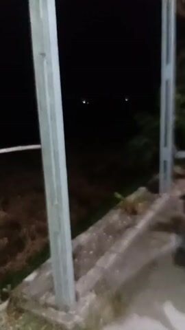 This is a vertical video taken at night, likely from a covered outdoor area. Two bright, reflective dots are visible in the dark background, suggesting the eyes of an animal. In the foreground, two vertical metal poles support a roof, and a motorcycle is parked on the right side. The ground appears to be a mix of concrete and dirt. The overall atmosphere is dark and quiet.