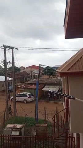 The video presents an aerial view of a bustling neighborhood street, likely in Narayi, Nigeria, under an overcast sky. The scene is dominated by dirt roads and numerous single-story buildings with corrugated metal roofs, suggesting a mix of residential and small commercial establishments.

Several utility poles with tangled power lines run across the frame, indicating the electrical infrastructure of the area. A rusty metal fence with barbed wire separates the foreground from the street. On the right, part of a building with a red-tiled roof and a dark brown spiral staircase is visible, leading up to an unseen level.

People can be seen walking along the dirt road and near the small shops. A white station wagon is parked on the road, and a motorcycle is parked near one of the buildings. The overall impression is one of everyday life in a lively community. In the background, a voice can be heard speaking a local language, possibly Yoruba, adding an auditory element to the visual scene. There is no visible text in the video.