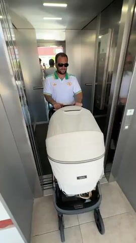 A man with sunglasses and a short beard wearing a white polo with green and yellow stripes around the sleeves and collar pushes a baby stroller into an elevator. He has tattoos on his left arm. Another woman is in the elevator, using her phone. The man then presses the elevator button. The elevator appears to be inside a building, with people visible outside the open doors. The interior of the elevator is stainless steel. The baby stroller is light-colored with a black base.