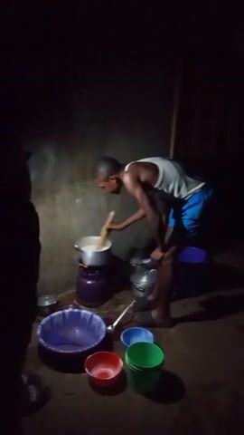 In a dimly lit room, a slender man wearing a white tank top and blue shorts is intently stirring a pot on a portable stove. He is leaning over the pot, with his left hand resting on his left knee for support. Various bowls in blue, purple, red, and green are scattered on the floor around him, along with other cooking utensils. To the left of the frame, a dark silhouette of another person is visible. The wall behind the man appears to be a painted concrete surface.