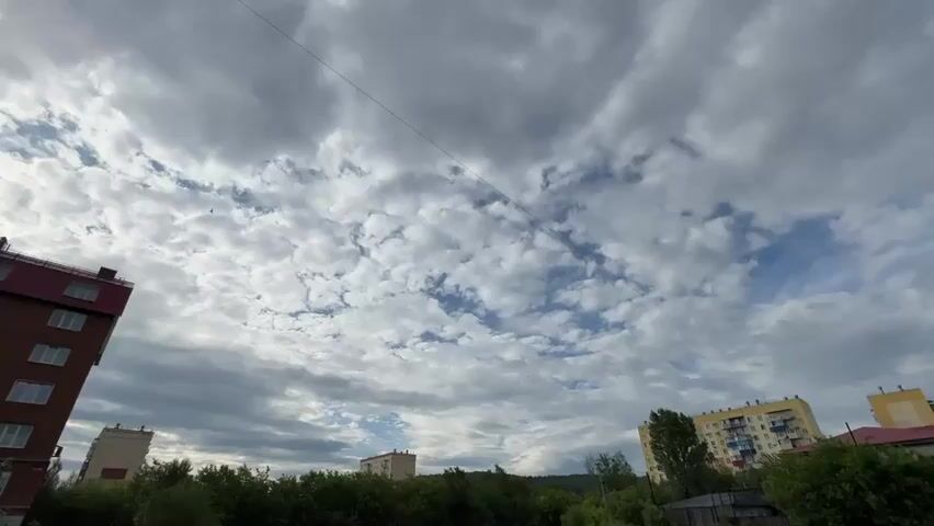 The video shows a cityscape with buildings and trees under a cloudy sky. The camera is pointed upwards, capturing the dynamic cloud formations. Two apartment buildings, one brick and one yellow, are visible on the right and left sides of the frame, respectively. Green trees and foliage form a dense line in the foreground, separating the buildings from the sky. A thin wire stretches across the sky from the top left to the top right corner of the frame. The lighting suggests it's daytime, possibly late afternoon or early evening, given the diffused light from the overcast sky. The overall atmosphere is serene and somewhat dramatic due to the cloudscape.