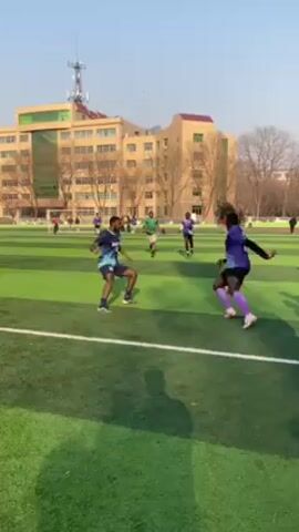 The video shows a group of people playing soccer on a green artificial turf field. They are wearing soccer jerseys of different colors: light blue, purple, and green. A tall building stands in the background with trees. The sky is clear and blue. The camera is hand-held and follows the action on the field, focusing on the players as they dribble, pass, and defend. A few people stand along the perimeter of the field, watching the game.