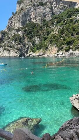 The video shows a bay with clear turquoise water, bordered by a rocky cliff covered with greenery. A person is swimming in the water. A small boat is anchored further away. A safety line with yellow floats is strung across the water, separating swimmers from the open sea. The sky is blue. The shot is taken from a slightly elevated position on the shore, with rocks in the foreground.