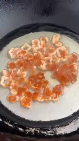 The video shows food frying in a large, wok-like pan. The food items are small, round shapes with a hole in the center and appear to be white and orange in color. Bubbles can be seen in the oil as it fries, and some steam rises from the hot pan. The pan itself looks worn, with a slightly stained or tarnished metallic surface.  The overall setting appears to be a kitchen or food preparation area.