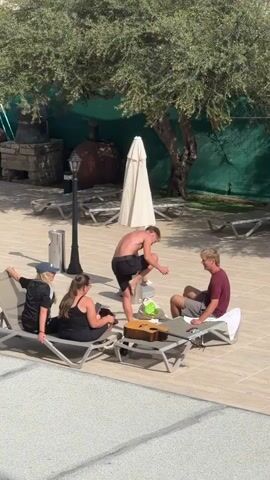 Here's a description of the video:

The video shows a group of four young people gathered on a tiled area near a swimming pool, likely a resort or hotel setting. Two of the people, both women, are sitting on a lounge chair. One wears a black shirt with white stripes and a baseball cap, the other is in a black tank top. Two men are also present; one is sitting on another lounge chair wearing a maroon shirt and gray shorts, while the other, shirtless and wearing dark shorts, initially stands and then walks away. A guitar case rests on a low table in front of the lounge chairs. An umbrella is closed nearby. The background includes an olive tree providing shade, other empty lounge chairs, a lamp post, and some stone structures. The scene appears sunny and relaxed.
