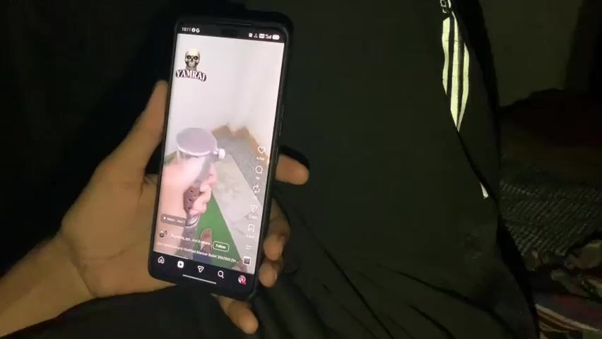 A person's hand holds a smartphone displaying a short video. The video shows a close-up of a metal cylindrical object with a cap, and the hand is turning the cap. The background of the video is a light-colored wall. The smartphone screen also shows social media interface elements, including a username "YAMRAJ," follower counts, and like buttons. The overall scene is dimly lit, suggesting an indoor environment, possibly at night. The person holding the phone is wearing dark clothing, including what appears to be black pants with white stripes.