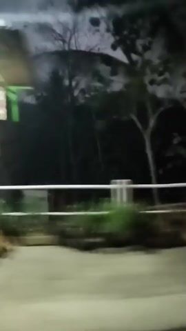 The video begins with a blurry shot of an outdoor seating area with dark chairs and an orange building in the background. The camera then pans to the left, showing a residential street at night. There are houses and trees lining the road, and the street is illuminated by streetlights and the headlights of a vehicle. The camera continues to move forward, showing more of the street and the surrounding houses. The overall impression is of a quiet, rural setting at night.