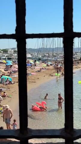 The video shows a crowded beach scene viewed through a metal grate. The beach is filled with people sunbathing, swimming, and relaxing. There are many beach umbrellas and towels spread out on the sand. In the water, some people are swimming or floating on inflatable toys, including a prominent pink flamingo float. A few people are standing in the shallow water near the shore. The background shows a harbor with numerous sailboats docked. The sky is clear and blue. The overall atmosphere appears to be a sunny, busy day at the beach.