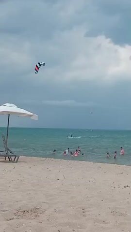 The video shows a beach scene at Shkorpilovtsi, Bulgaria. The sky is cloudy, suggesting it might be an overcast day or that a storm is approaching. The sea is a vibrant turquoise color, with some waves.

In the foreground, a sandy beach is visible, with two empty lounge chairs and a white umbrella. Several people are in the shallow water, appearing to be swimming or wading. Some of them are children, while others are adults.

Further out in the sea, there are a few individuals engaged in watersports. A prominent kite surfer with a black, white, and red striped kite is visible in the middle ground, actively riding on the water. Another kite surfer is further in the distance on the right. The presence of kite surfers suggests windy conditions suitable for this activity.

The overall mood of the scene is relaxed and leisurely, with people enjoying the beach and the water. The cloudy sky adds a dramatic element to the otherwise idyllic beach setting. There is no visible text in the video.