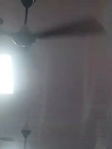 A ceiling fan is rotating in a room. There is a bright light source on the left side of the frame. The fan blades are blurred due to their motion. The room appears to be dimly lit, with the exception of the bright light source. The color of the ceiling is a light gray.