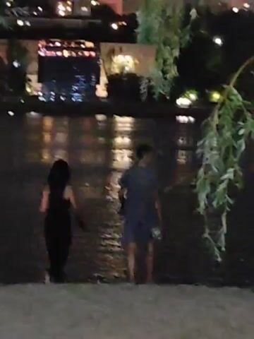 The video shows two people standing at the edge of a body of water at night. The person on the left is wearing a dark dress and the person on the right is wearing a dark shirt and shorts. The water reflects the lights of buildings in the background. A tree branch with green leaves hangs into the frame from the right side. The people appear to be looking out at the water.