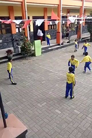 The video shows an outdoor courtyard area with a brick-paved surface. Several children, mostly wearing yellow shirts with blue pants, are present. Some children appear to be playing a game, possibly volleyball, with one child in the foreground looking towards the center of the activity. In the background, there is a building with orange and yellow accents, decorated with red and white triangular flags. Several adults are also visible in the background, some standing near the building entrance. There are trees, plants, and a motorcycle parked to the right of the frame. The overall scene suggests a schoolyard during some kind of event or activity.