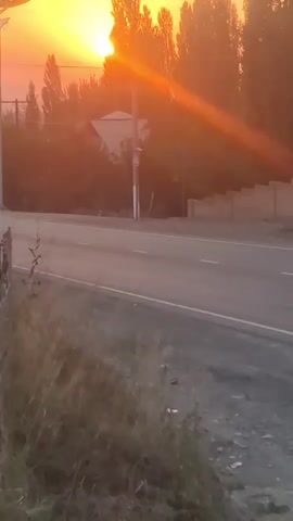 The video shows a sunset over a rural road. The sun is setting behind a line of trees, casting a warm orange glow over the scene. A road runs through the middle of the frame, with dry grass and dirt on either side. There are a few buildings in the background, and a street light. The camera is stationary, and the video is shaky, suggesting it was filmed by hand. There is no sound.