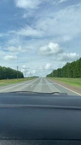 A dashboard view from inside a car shows a straight, paved road stretching into the distance under a partly cloudy blue sky. The road is lined with trees on both sides. The road surface appears uneven with alternating lighter and darker colored patches forming stripes. Several utility poles are visible along the left side of the road. A white car is visible in the distance on the road.