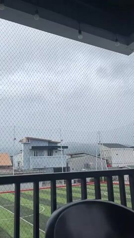 The video pans across a view of a residential area under an overcast sky. In the foreground, a black chair is partially visible. Beyond the chair is a black railing. Through the railing and a fine mesh net, a green artificial turf field with white markings is visible, suggesting it might be a sports field. Several houses are seen in the midground, varying in color and architectural style. Some have flat roofs, while others have angled ones. In the background, faint outlines of mountains are discernible through the hazy atmosphere. The overall impression is a residential neighborhood on a cloudy or potentially rainy day.