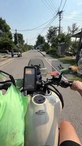 This video captures a motorcycle ride on a sunny day, likely in Kandi, India. The camera is mounted on the handlebars of a cream-colored Royal Enfield Himalayan motorcycle, offering a first-person perspective. A bright green plastic bag is secured to the left side of the handlebars. Prayer flags, displaying colors and patterns, are attached to the motorcycle's mirrors and handlebars, suggesting a spiritual or cultural element to the journey.

The scene is a paved road lined with lush green trees and vegetation on either side. In the distance, power lines stretch across the sky, and a few buildings are visible. Other vehicles, including a scooter and a motorcycle with riders, are present on the road, indicating moderate traffic.

The prominent subjects are the two individuals on the motorcycle. The rider in the foreground, wearing a black t-shirt and a helmet, is looking directly at the camera with an open mouth, appearing excited or surprised. Visible text on his t-shirt reads "СВОБОДНЫЙ СВОБОДНО". He is wearing a silver watch on his left wrist. The passenger behind him wears sunglasses and a beige jacket, looking ahead. Their expressions suggest an enjoyable and possibly adventurous outing.

The time of day appears to be daytime, given the bright sunlight and clear blue sky with some clouds. The weather seems pleasant for riding. The overall atmosphere is one of a scenic road trip, possibly through a rural or semi-urban area in India, characterized by the presence of natural beauty and local cultural elements.