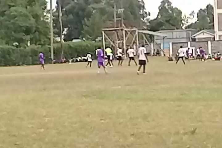The video shows a soccer game on a grassy field. Players in white and purple jerseys are scattered across the field, seemingly engaged in a match. Some players are running, while others are standing or walking. On the sidelines, a group of people sit and watch the game. The background features trees, a building, and a wooden structure, potentially a goal or support for netting. The sky appears overcast, suggesting a cloudy day.