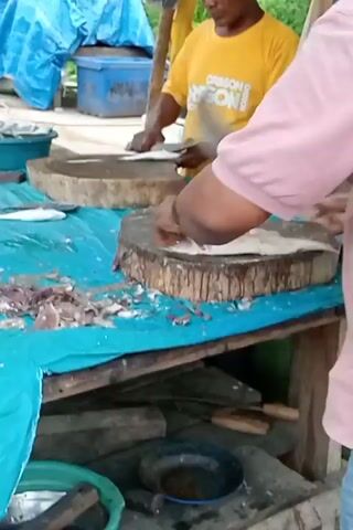 The video captures two men engaged in cleaning and processing fish at an outdoor setup, likely a market stall in Pulorayeuk, Indonesia.

In the foreground, a man wearing a light pink short-sleeved collared shirt and blue jeans is seen from behind, intensely focused on his task. He uses a large cleaver to chop or clean fish on a thick, circular wooden chopping block. Fish scales, guts, and other discarded parts are visible on the blue tarp covering the wooden table surface around his work area.

Another man, wearing a yellow short-sleeved t-shirt with some faded text (partially visible, possibly "OREGON"), stands further back and to the left. He is also actively preparing fish on a similar wooden chopping block, using a knife. He appears to be scaling or gutting fish.

The scene is rustic, with a simple wooden table structure draped with a bright blue tarp. On a lower shelf of the table, a black pan or bowl is visible. A green bowl containing fish rests on the ground next to the table. The background shows more blue tarp or plastic sheeting, possibly covering goods or part of a makeshift shelter, and a blue plastic container. The ground appears to be uneven earth. The lighting suggests it is daytime, and the men exhibit focused and diligent expressions as they work.