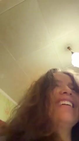 This video appears to be a selfie-style recording of a woman with long, curly brown hair. She is indoors, and the ceiling with a light fixture is visible. The woman is laughing and moving her head. In one brief moment, another person, an older woman with short gray hair and glasses, is visible in the foreground. The younger woman then turns back towards the camera and hugs the other person. The younger woman is wearing a bracelet and a ring. The background includes what looks like wallpaper and possibly a piece of furniture.