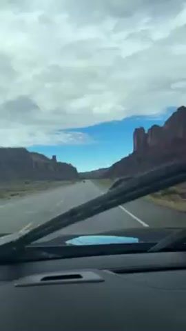 The view from inside a car shows a straight, empty road running through a desert landscape. On either side of the road are large, reddish-brown rock formations resembling cliffs or buttes. The sky is blue with scattered white clouds. The car's windshield has been recently cleaned, leaving streaks of water on the glass, and a windshield wiper is visible in the foreground. Other cars are seen in the distance on the road ahead.