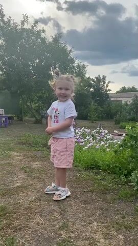 This is a short video clip, featuring a young girl standing in an outdoor setting. She is the sole human subject. The girl is wearing a white t-shirt with a graphic print, pink shorts, and sandals. Her hair is styled in two small buns. She appears to be looking slightly off-camera with an expression of surprise or excitement, her mouth open.

The scene is a grassy yard with dry patches of ground. In the background, there are trees, green foliage, and a flower bed with white and purple flowers. The weather appears to be overcast with a cloudy sky. The light suggests it is daytime, possibly late afternoon or early evening. There are hints of structures in the distance, possibly a building or fence, and some objects like a purple stool or chair are visible on the left side of the frame.

The activity taking place is minimal; the girl is standing still, but her posture and expression suggest she is reacting to something outside the frame or has just been addressed. The overall atmosphere is casual and domestic, indicative of a backyard or garden space. No visible text is present in the video. The location context provided is Маринопіль, Ukraine.