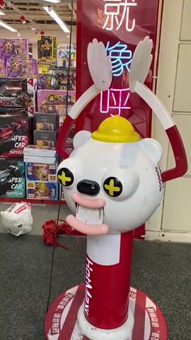 A short video clip shows a playful, cartoonish white bear-like figure with red arms raised above its head, wearing a yellow hard hat. The figure has large, cartoonish eyes with yellow crosses and two pink sausage-like shapes for a mouth. It stands in front of a red sign with Chinese characters in neon lighting. In the background, shelves are filled with toy cars and other toys. The floor is grey with red and white striped accents around the base of the figure. A white plastic bag and some red material are on the floor to the left of the figure. The lighting is bright, suggesting an indoor commercial setting.