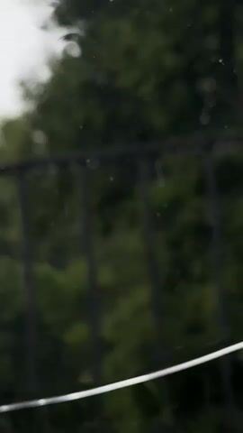 This is a close-up, out-of-focus video of raindrops on a window. The background is blurred green foliage with a dark railing visible.