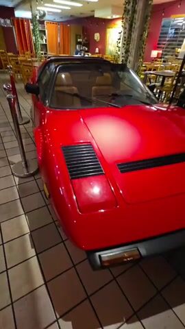 The video shows a red convertible sports car, possibly a Ferrari, parked indoors. The car is positioned at an angle, with its front to the right of the frame and its side facing the camera. The interior of the car appears to have tan leather seats. There is a "Please Do Not Touch" sign on the driver's side window. The car is cordoned off with stanchions and velvet ropes. The background suggests the car is in a restaurant or entertainment venue, with tables, chairs, and decorative elements like hanging plants and artwork visible. A mural depicting the Colosseum is present in the background. The lighting is artificial, creating a warm atmosphere.