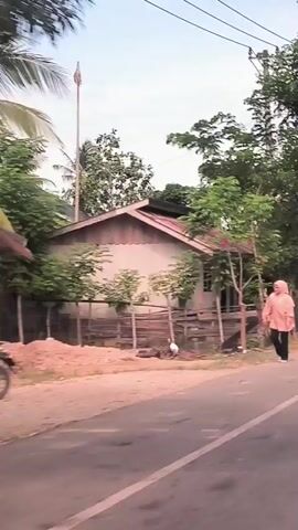 The video depicts a street scene in what appears to be a rural area. The camera is likely mounted inside a moving vehicle, giving the perspective of someone driving along the road. Initially, a person wearing a peach-colored hooded jacket and dark pants is seen walking alongside a building with a rusty roof. As the vehicle advances, groups of people dressed in traditional attire including hijabs, are seen walking along the road. Some are carrying bags. The road is paved with a white line marking its edge. Trees with dense foliage line the side of the road, casting shadows. Some smaller buildings and houses are visible. As the camera moves, motorcycles pass by on the road.