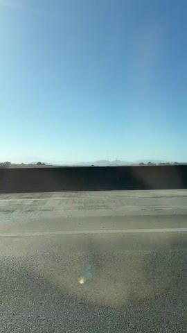 A static, vertical shot of a highway from inside a vehicle. A dark, concrete barrier runs horizontally across the middle ground. The sky is a clear blue with the sun visible in the upper left corner. Distant hills and some trees are visible beyond the barrier. The road surface in the foreground is asphalt and appears to be in good condition. There are no people or other vehicles clearly visible in the frame.