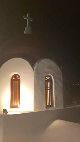 The video shows a nighttime exterior view of a white building with a domed roof and a cross at the apex. There are two arched windows with stained glass that are illuminated from within. The building appears to be a church or chapel. The lighting is dim, and the sky is dark. There is a faint glow in the distance, suggesting a town or city. The camera is positioned slightly below the building, looking up. There is a sense of mist or fog in the foreground.