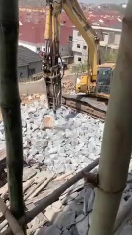 From a vantage point on a bamboo structure, a yellow KOMATSU excavator with a hydraulic breaker attachment is seen demolishing a pile of gray rocks.  The breaker repeatedly impacts the rocks, generating dust and small chips.  In the background, there are houses with red-tiled roofs. A person can be seen operating the excavator from within the cab.  The sky is clear and blue.