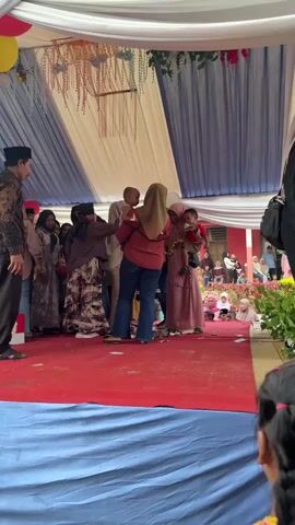 The video shows a gathering of people, likely at an outdoor event with a tent structure overhead. Several individuals are visible in the foreground and midground. On a raised red carpeted area, a woman in a reddish-brown top and blue jeans holds a baby. Another woman in a light pink headscarf and dress stands next to her. To their left, a group of people are gathered, some wearing headscarves and traditional clothing. In the midground, a red building with white trim is visible, and a crowd of people are seated and standing in front of it. There are also green trees and foliage in the background. Several balloons in pink, red, and yellow are attached to the tent structure. In the lower right corner, the back of a person with dark hair in pigtails is visible, facing away from the camera. The overall atmosphere appears to be festive.