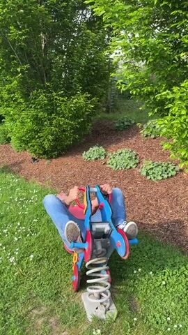 A man is sitting on a spring rider toy in a park. He is wearing a red t-shirt, blue jeans, and white sneakers. The spring rider toy is blue, red, and yellow, and it is mounted on a gray spring. The man is smiling and appears to be enjoying himself as he rocks back and forth on the toy. The background includes green grass, trees, and wood mulch. The scene is well-lit.